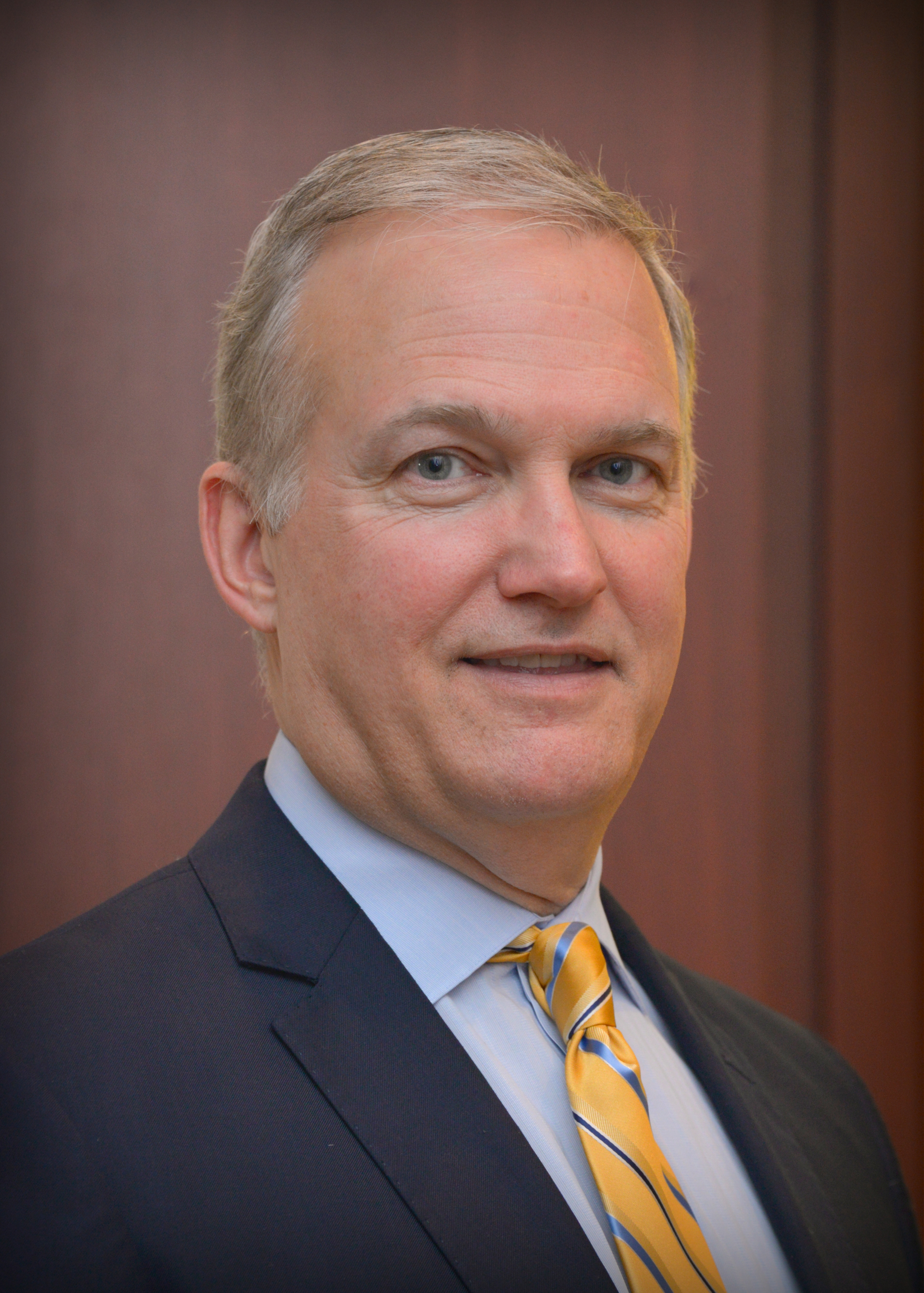 Headshot of BIll Ellis with his short, grey hair parted to the side wearing a navy blazer with a light blue shirt, and a yellow tie with a blue stripe.